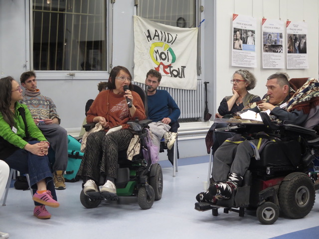 Une photo de la soirée handimoitout : une personne en fauteuil roulant prend la parole lors d'un échange. A l'arrière-plan, la bannière d'handimoitout.